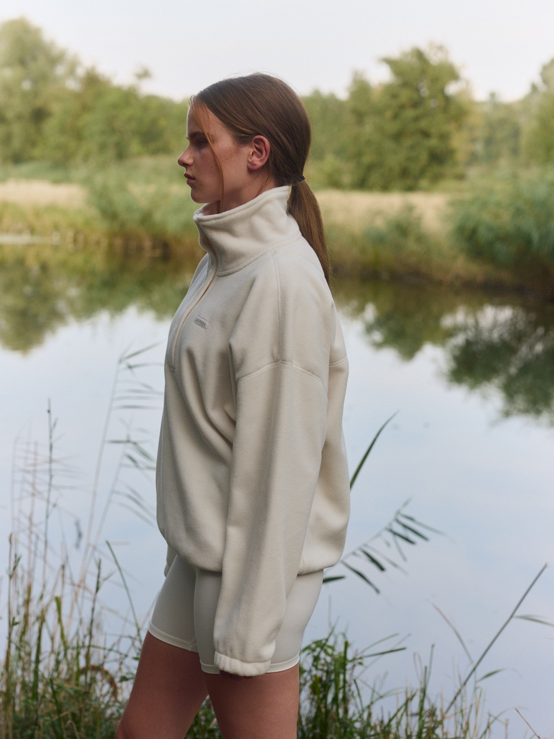 Woman wearing a creme polar fleece half-snap pullover standing outdoors by a lake.